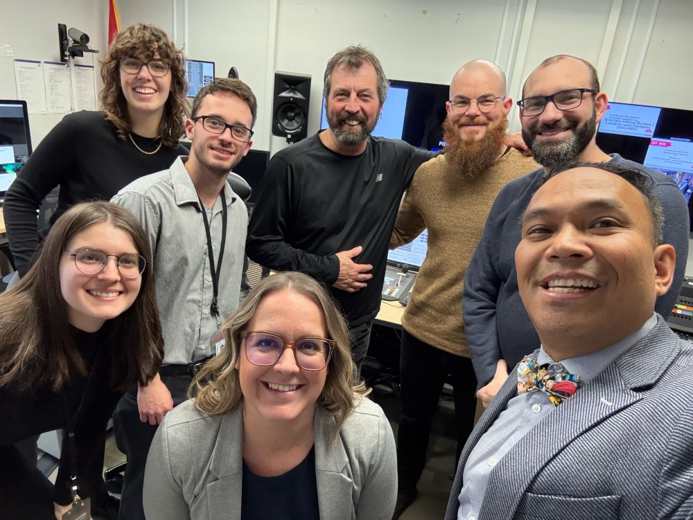 A group of eight smiling individuals gathered in what appears to be a control room. They are surrounded by monitors, speakers, and technical equipment, suggesting a media or production environment. The group includes a mix of men and women, with the person in the foreground on the right wearing a distinctive bow tie and suit, possibly the host or presenter. Everyone appears friendly and engaged, reflecting a collaborative team dynamic.