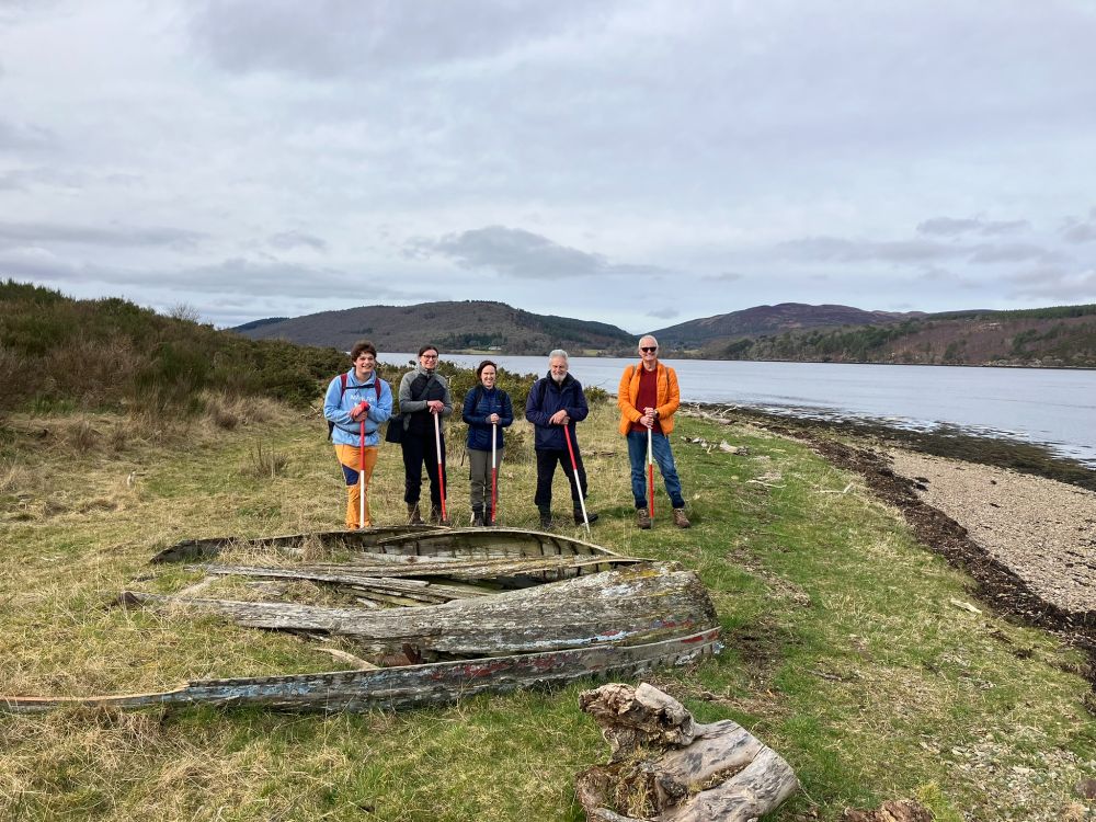 A group of five SCAPE volunteers stand next to an old, ruined fishing boat on the foreshore of the Dornoch Firth.