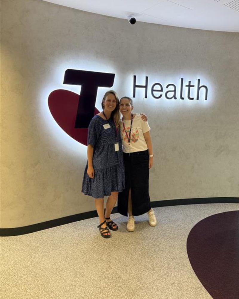Two smiling women stand together in front of a wall displaying the illuminated Telstra Health logo. Both wear name badges and lanyards, posing in a bright, modern office foyer.