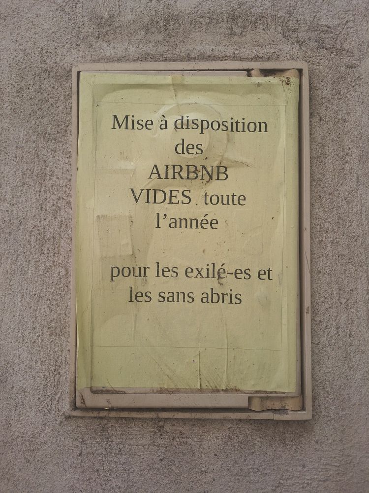 Affichette collée sur un mur. Il est écrit "Isère à disposition des Airbnb vides toute l'année pour les exilé-es et les sans abris