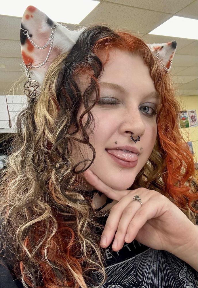 Selfie of CandiedCalico while working at local game shop. Curly calico dyed hair and wearing calico cat ears with pentacle chain earrings. She also has fang piercings and a black spiked septum ring. She smiling with her tongue sticking out in a mischievous expression. 
