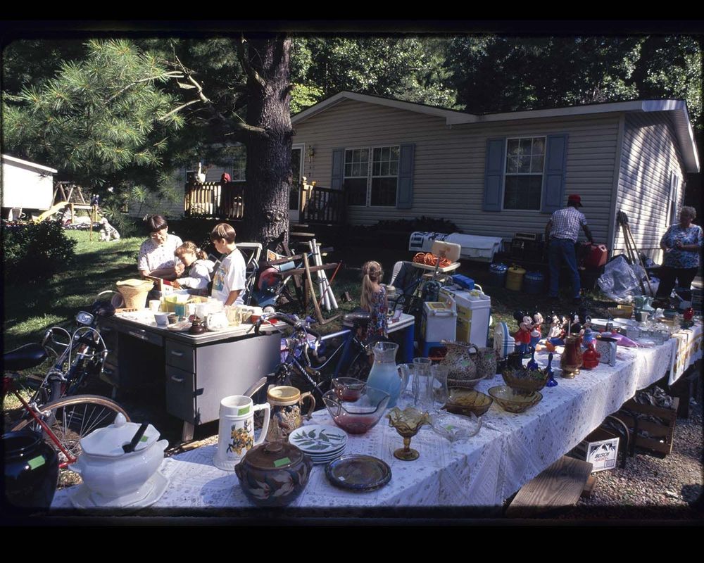 Marilyn Moore & 2 children browse books amongst furniture, toys, glassware, and assorted housewares at backyard yard sale.