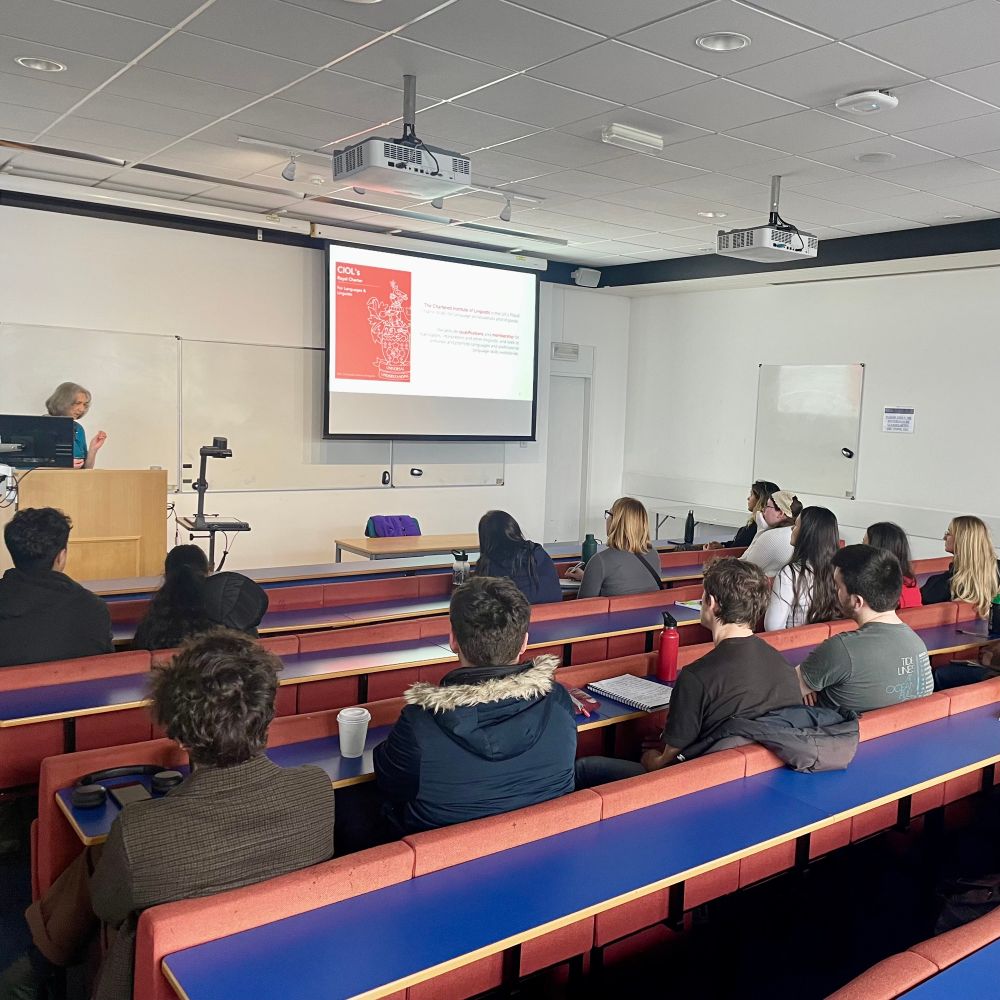 A lecture theatre (a room for teaching where the seating arrangement is tiered,) with students looking on towards the front of the room, towards a speaker (a woman) only the back of the students can be seen, as they face forward. It’s daytime.
