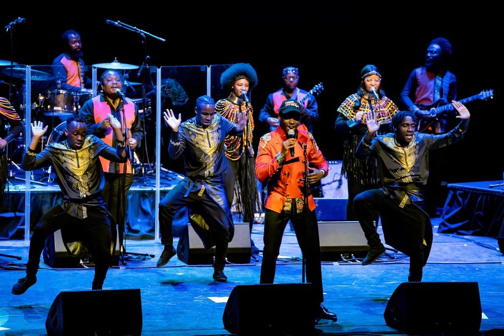 A band performing with vocalists and dancers on stage.