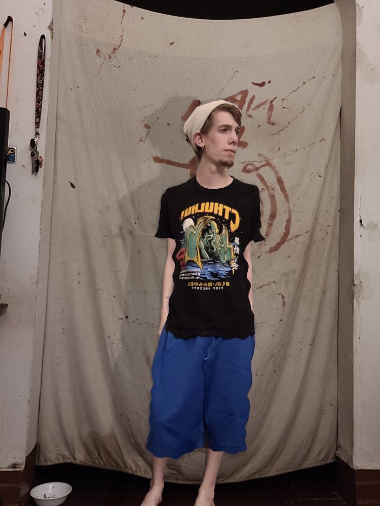 a man, with facial hair stands, looking to his left. He's wearing a beanie, a black cthulhu t-shirt and blue shorts. barefoot. he's slightly shorter than the previous photo. Part 2 of a height shrinking sequence.