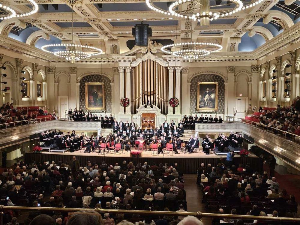 Large well lit concert hall, soaring pipe organ behind the stage. Chorus and Orchestra seated on stage. Audience seated on flior and balconies.