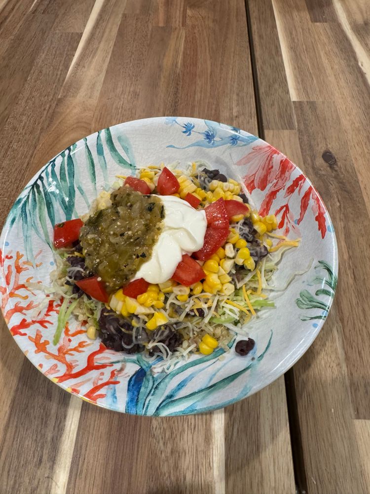 A bowl with quinoa, lettuce, black beans, corn, tomato’s, shredded cheese, sour cream, and salsa verde