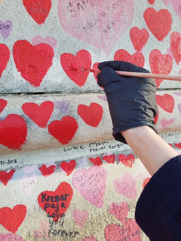A black-gloved hand refreshes the red paint on one of the hearts on the memorial wall.