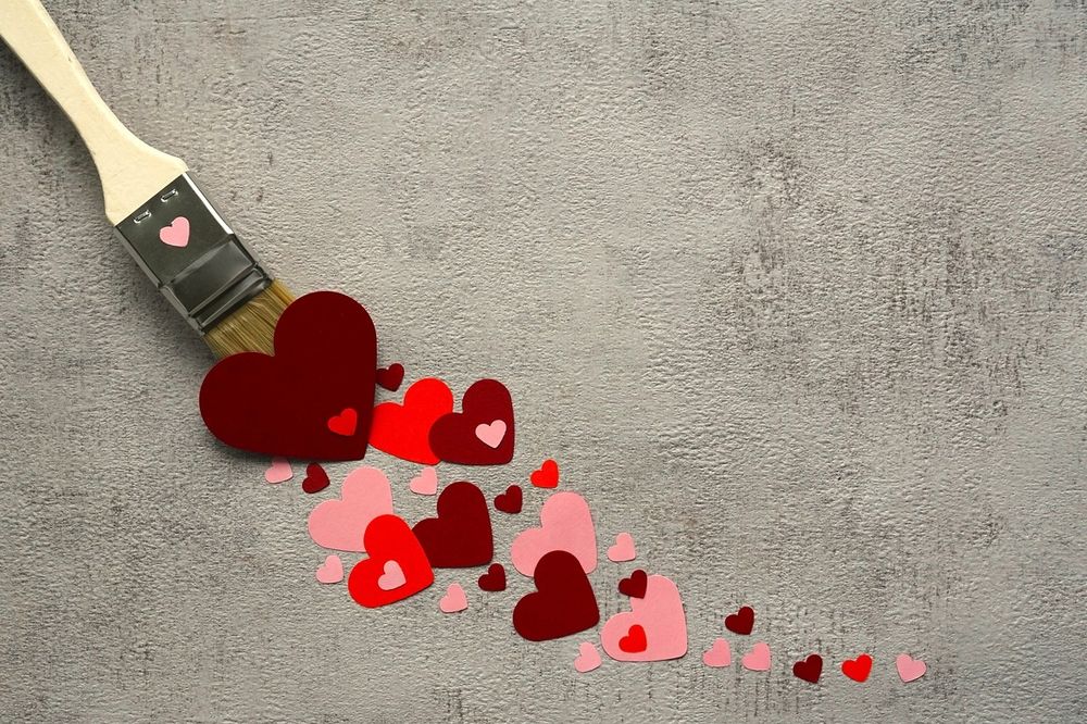 A paintbrush being pulled along a concrete wall, leaving red, burgundy, and pink hearts in its wake.