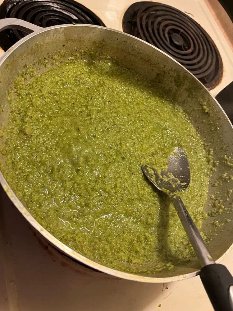 Photo of a caldero filled with a rich green sofrito. A mixing spoon is sitting in the caldero and the caldero is resting on top of an electric stove.