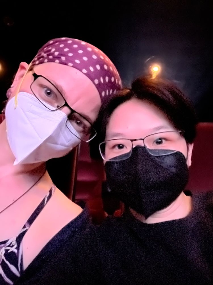 A selfie of two people one wearing n-95 white mask other wearing of-94 black mask in a movie theater.