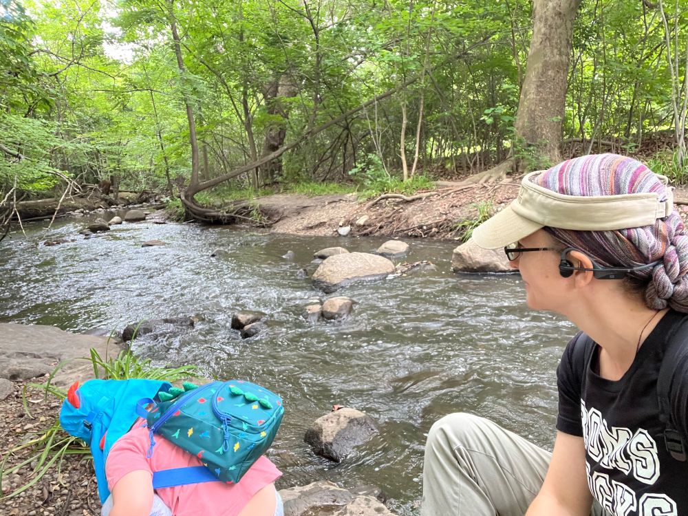 A toddler and an adult sit on the bank of a river. The toddler is leaning to grab rocks to continue hurling them into the river. The adult is watching the toddler within arms length just in case and enjoying the sights and sounds of the environment. 