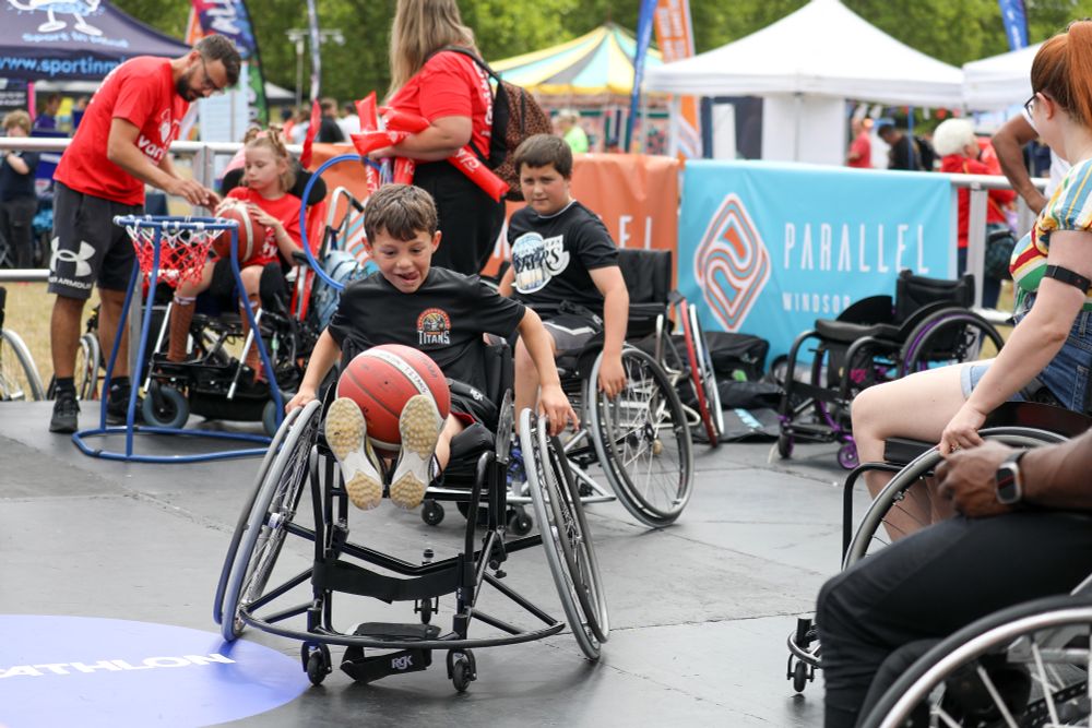 A photo in the Parallel Actice lifestyle zone which has multiple people on sport wheelchairs and a basketball ring on the left. the focus of the image is on a little boy sat in a sports chair with a black 'titans basketball' t shirt on and a basketball on his knees. His tongue is stuck out the side as he concentrates with a big grin! In the background are other tents and activities. 