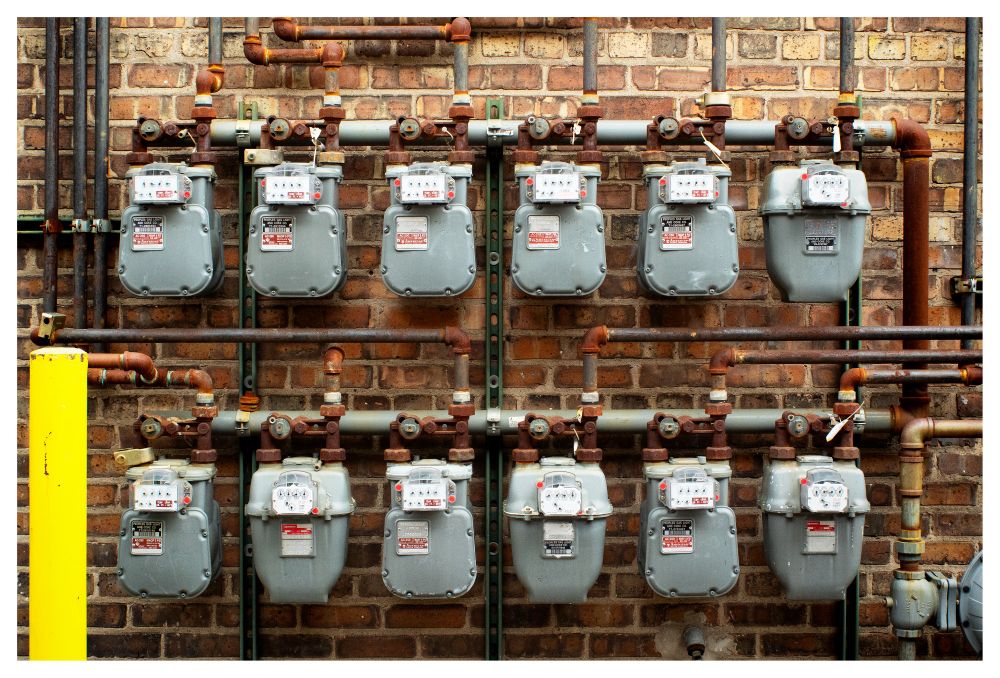 A color photograph of a series of gas meters and pipes affixed to a brick wall in an alley in Chicago's South loop. There is a bright yellow concrete pole just to the left.