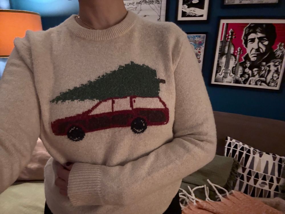 A person wearing a light beige sweater stands indoors, shown from the shoulders to the waist. The sweater features a large knitted design of a red station wagon carrying a green Christmas tree on its roof. The person’s left hand rests on their abdomen. Behind them is a dark blue wall decorated with framed artwork, and part of a sofa with patterned cushions is visible.