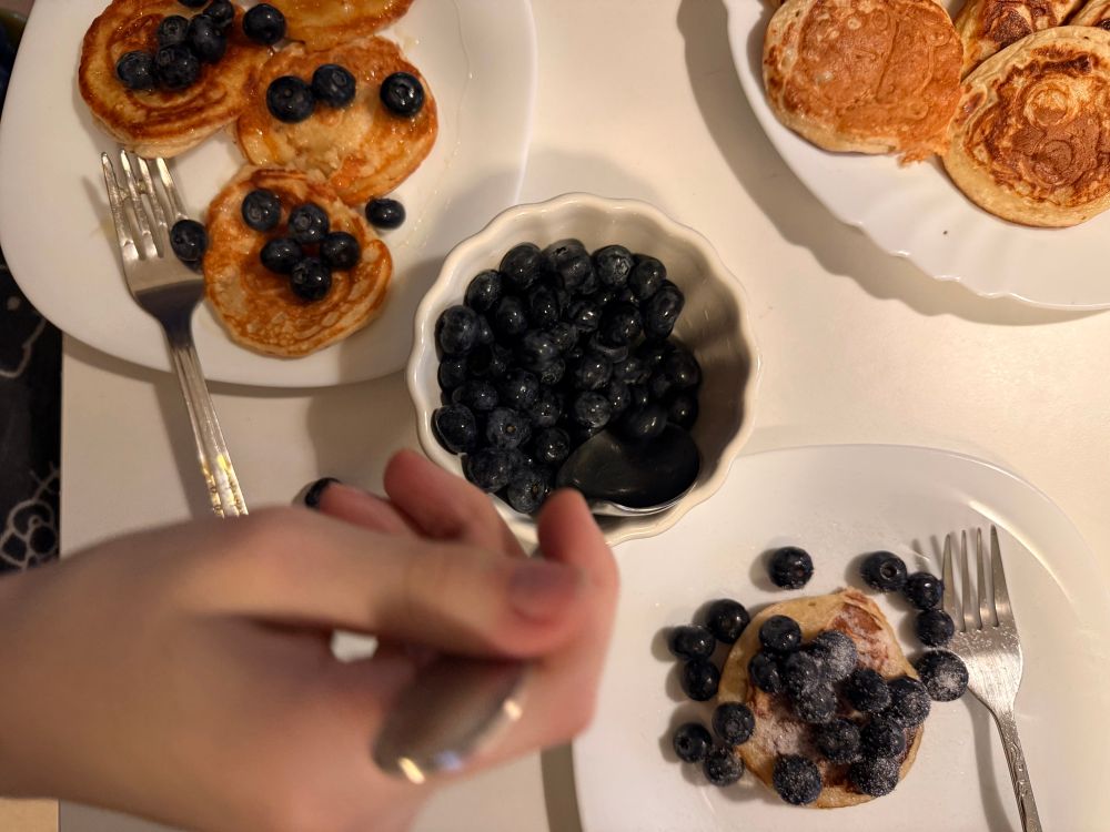 A table set for breakfast with several white plates of small, round pancakes topped with fresh blueberries. In the center is a scalloped white bowl filled with blueberries and a small spoon. A person’s hand holding a fork is visible in the foreground, reaching toward a plate with a single pancake covered in blueberries and powdered sugar. Two forks rest on the plates. 