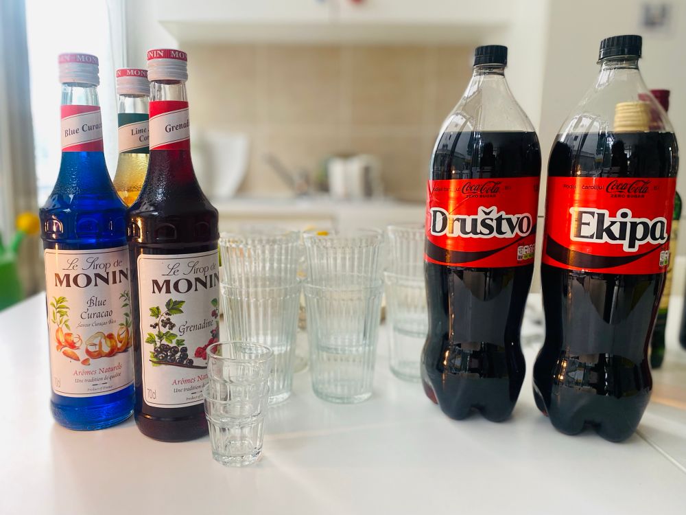 A kitchen table display with two Monin syrup bottles (Blue Curacao and Grenadine), a third syrup bottle partially visible (Lime Cordial), and two Coca-Cola bottles labeled “Društvo” and “Ekipa” in red and black branding. Several empty glass cups and shot glasses are arranged around the bottles in a brightly lit room.