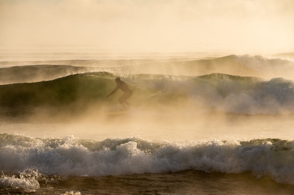 Surfer wearing a black wetsuit riding a 6' roller on Lake Superior on a -20f° morning in February 2022 during morning golden hour 