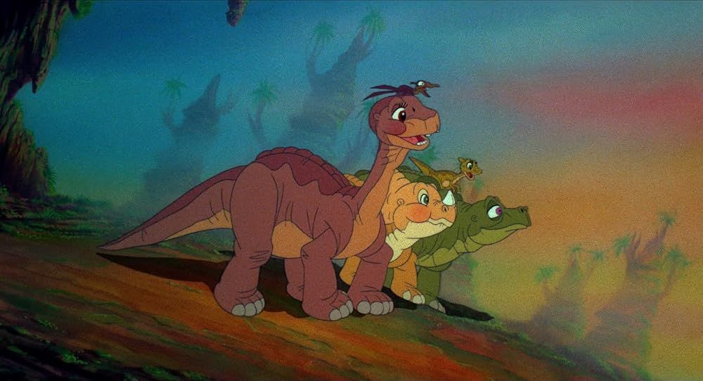 A cell image from the Land Before Time 1988 feature. It has all 5 of the dino babies in it, they are up on a smei grassy dirt hill overlooking a valley, it has streaks of red, purple, yellow, orange, green, and blue among the sky. making it appear a little foggy or "dreamy". Closest one is Littlefoot, with Pteri ontop of his head. Pteri's is a small brown type of Pteranadon. Next to them is Cera, a yellow blushfaced Triceratops / Ceratopsian. behind her is Spike, a green stegosaurus with Ducky on his back. Ducky is a small yellow-brown duck-billed dino (the odl website referred to her as a Parasaurolophus but she's likely closer to a Saurolophus.