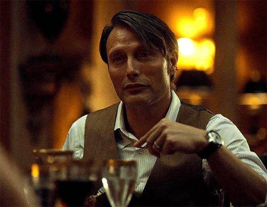 A small short gif of Mads Mikelsen as Hanibal Lecter from Hannibal. His arm is slightly bent and raised holding a fork, it's very candle-lit ambient lighting. He seems bemused, and begins talking, turning his head to the side to look over at someone else. There's some gold rimmed glasses infront of him