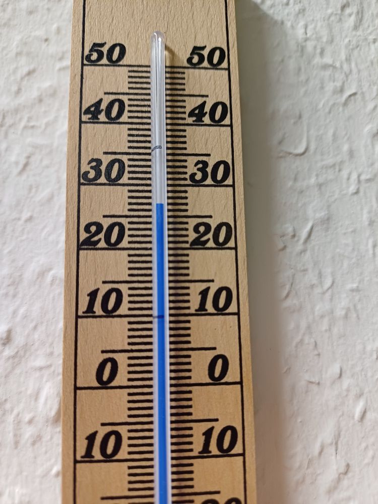 Bild eines Holzthermometers vor einer weißen Wand, die Anzeige steht bei knapp 27 ° C.