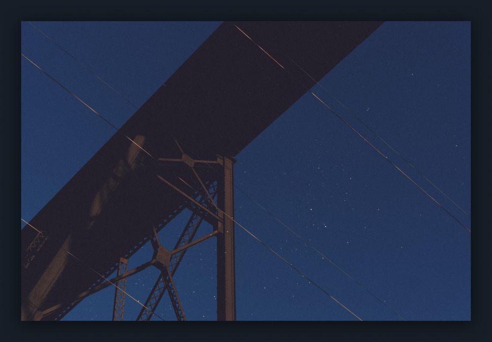 A photo looking up at a massive steel train bridge with electrical cables crossing the frame perpendicular to the bridge.
