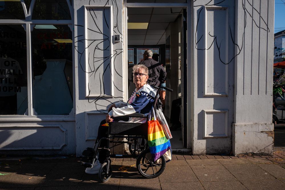 An older person in a wheelchair at Wirral Pride 2025 in New Brighton, holding a rainbow flag bag.
