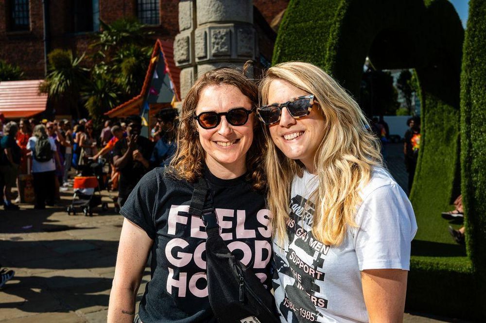 Two women smiling at the camera during Liverpool Pride. The woman on the left is wearing a black t-shirt with the words 'Feels Good Home' printed on it. The woman on the right is wearing a white t-shirt with text on it. In the background, there are other people, buildings, and greenery.