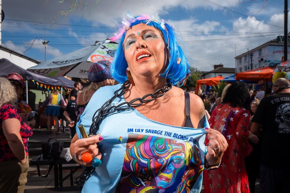 A person with blue hair and makeup is holding up their shirt at a Pride event. The shirt says 'IM LILY SAVAGE THE BLONDE BARS'.