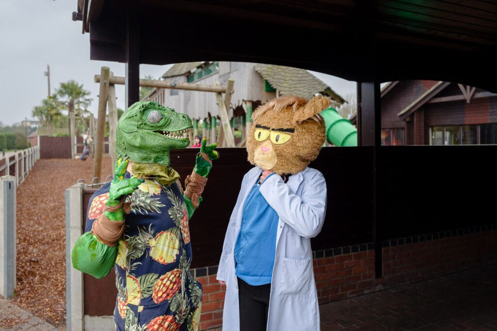 Two people in cosplay, one dressed as a dinosaur and the other as a cat, stand talking near a playground.