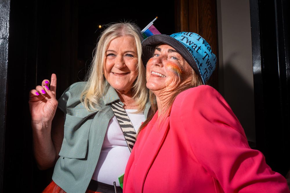 Two smiling women posing for a photo at Wirral Pride. One woman is wearing a hat with the word "Twat" printed on it, and the other is wearing a green jacket.