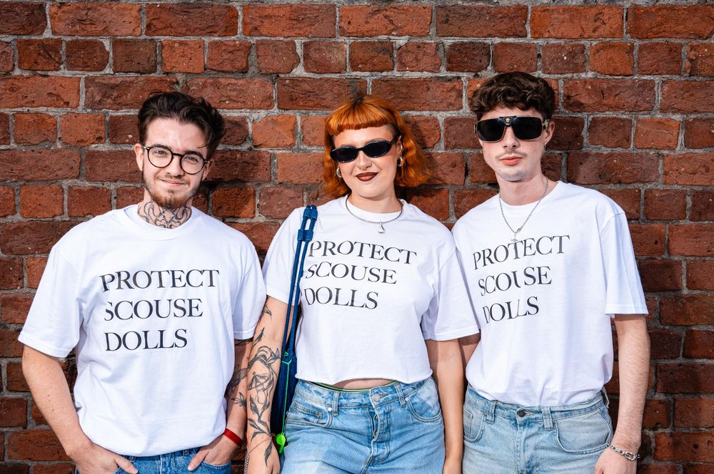 Three people wearing white t-shirts with the text "PROTECT SCOUSE DOLLS" standing in front of a brick wall. The image is taken during Liverpool Pride.