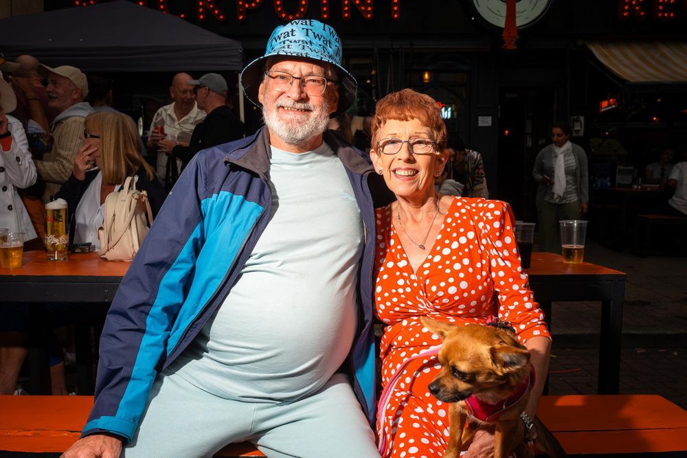A couple smiling at the camera, seated outdoors at a Pride event. The woman is holding a small dog.