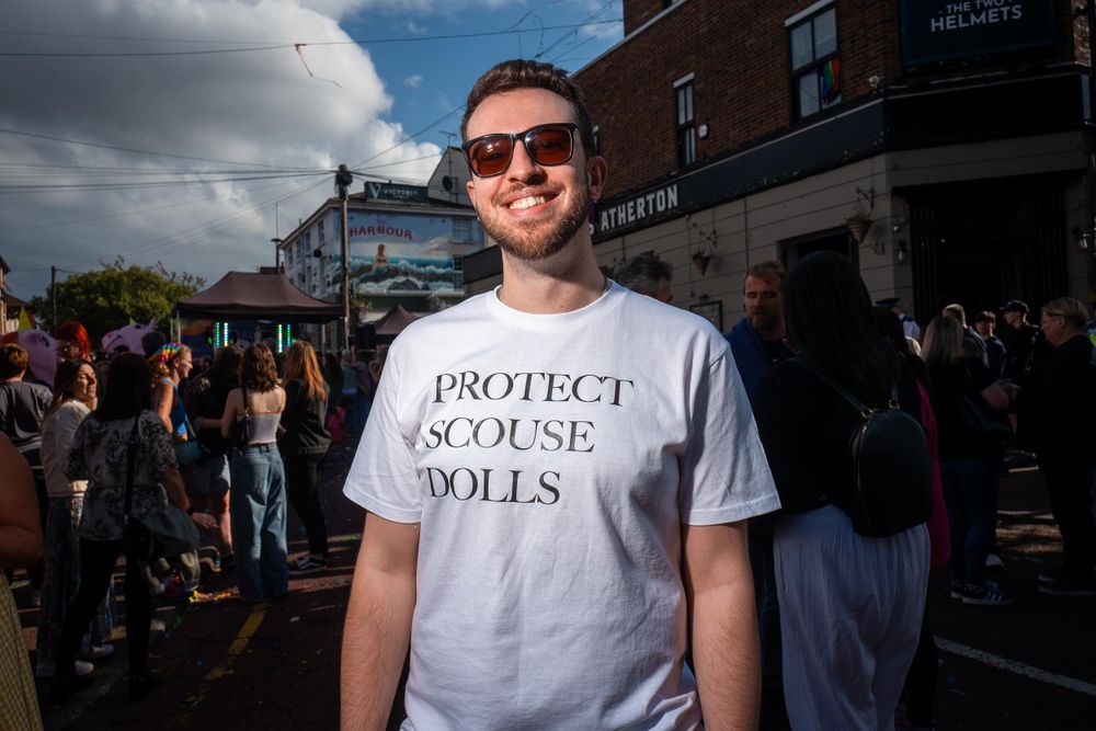 A man wearing sunglasses and a t-shirt that says 'PROTECT SCOUSE DOLLS' smiles at the camera in front of a crowd of people at Wirral Pride.