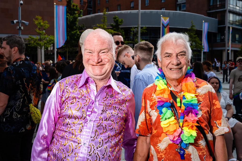 Two men smiling at the camera at Liverpool Pride, one wearing a pink sequined shirt and the other wearing a Hawaiian shirt and a rainbow lei.