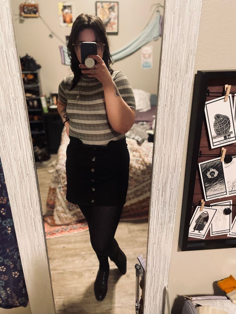 A selfie in front of a floor length mirror. I’m in my work outfit—a black a-line skirt, black tights, black oxfords, and a short sleeved top in olive green and beige. I’m posed with a hip popped, trying to look cute. Brown hair is partially pinned back, partially down. Behind me in the mirror, you can see my unmade bed and general bedroom messiness. 