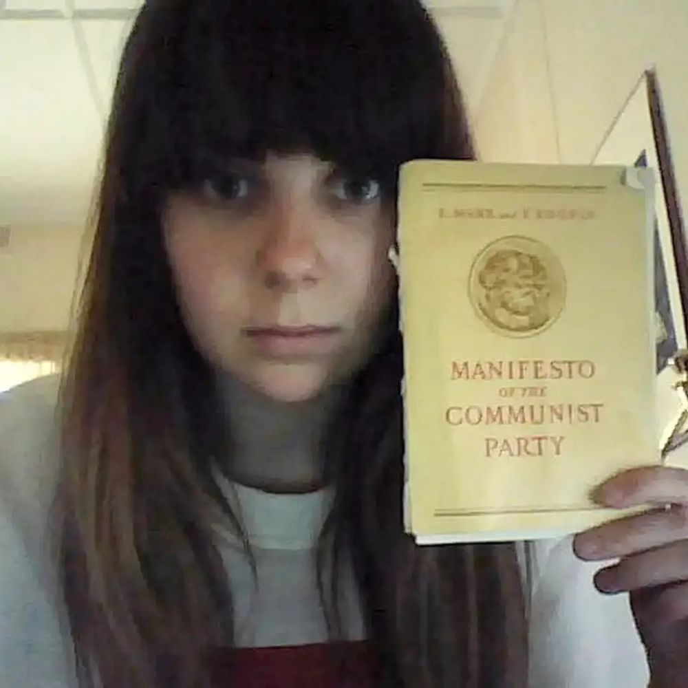 17-year-old me, with long brown hair and bangs, staring into the cameta holding the Manifesto of the Communist Party