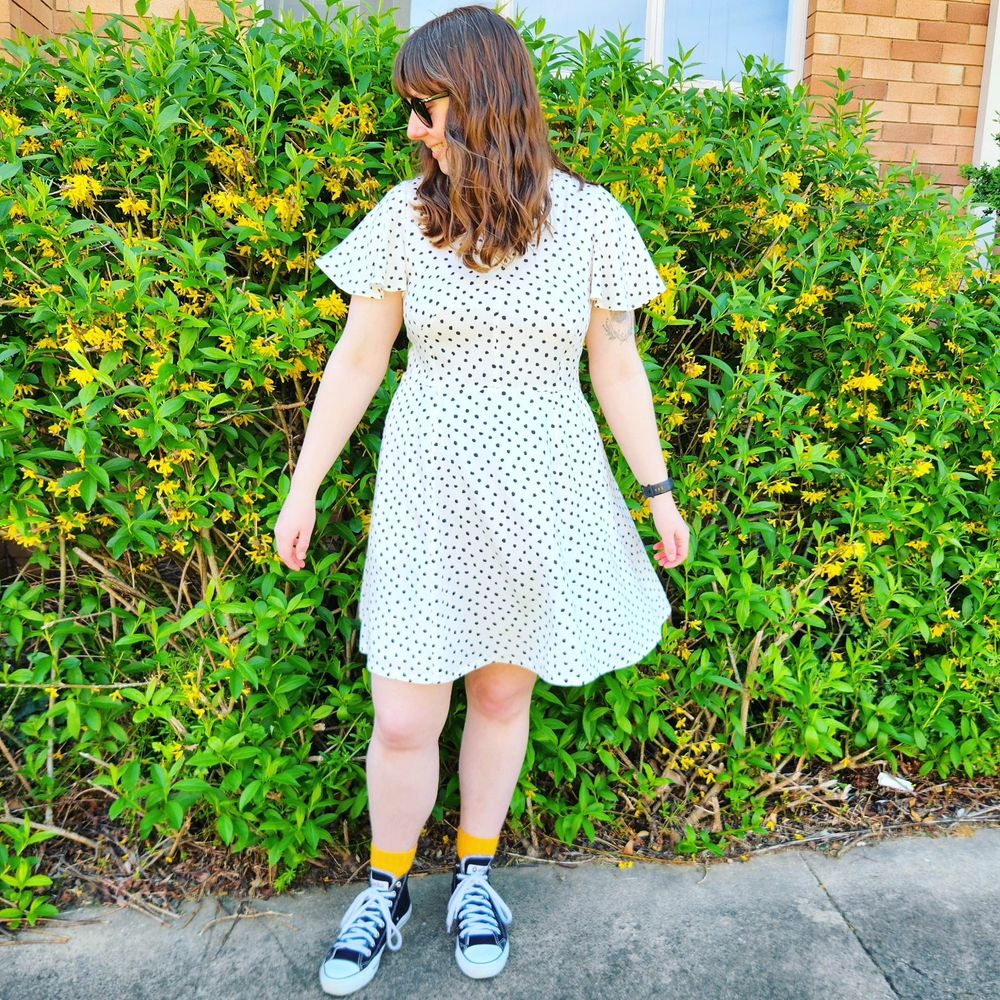 Me, a white woman with brown hair and bangs, in a cream and black handmade spotty dress and black converse and handknit yellow socks