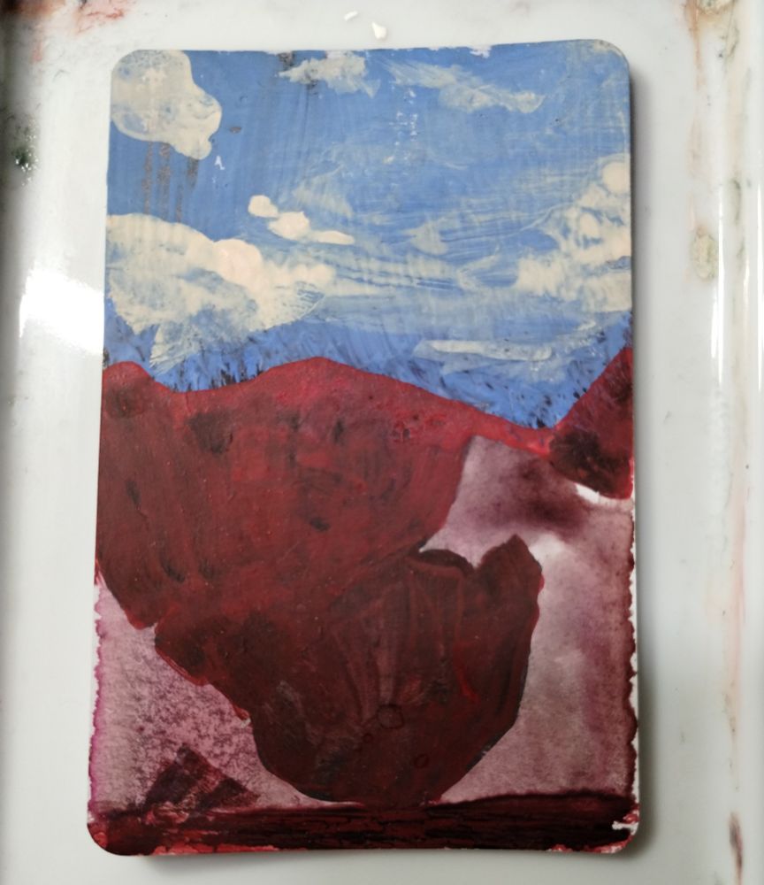 a small card painting of red mountains against blue skies. creamy painted clouds sit wispy over crimson ink forming the shadow shapes of peaks