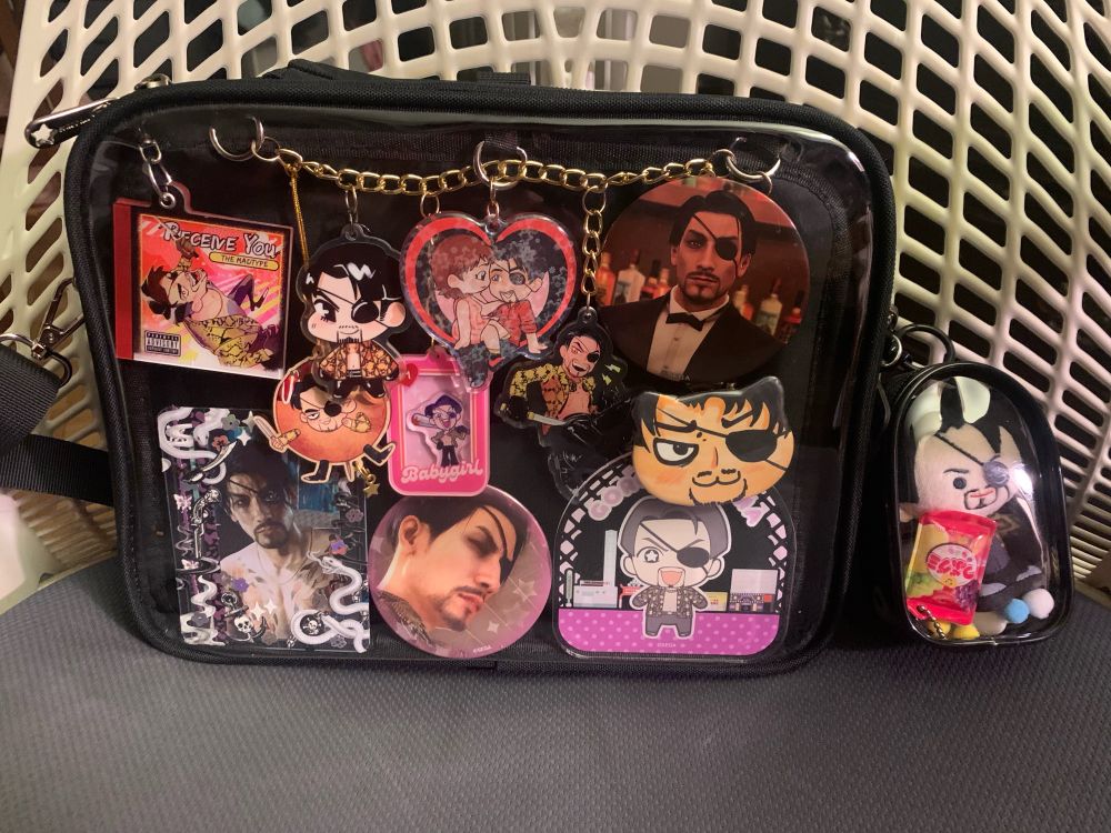 Picture of a black ita bag/purse on a chair. The ita bag has charms, buttons, and pictures of Goro Majima in it. Some are official merchandise, others are various artists or handmade pieces. There is also a small pouch attached to the bag that has a Kinokos mini Majima plush inside. 