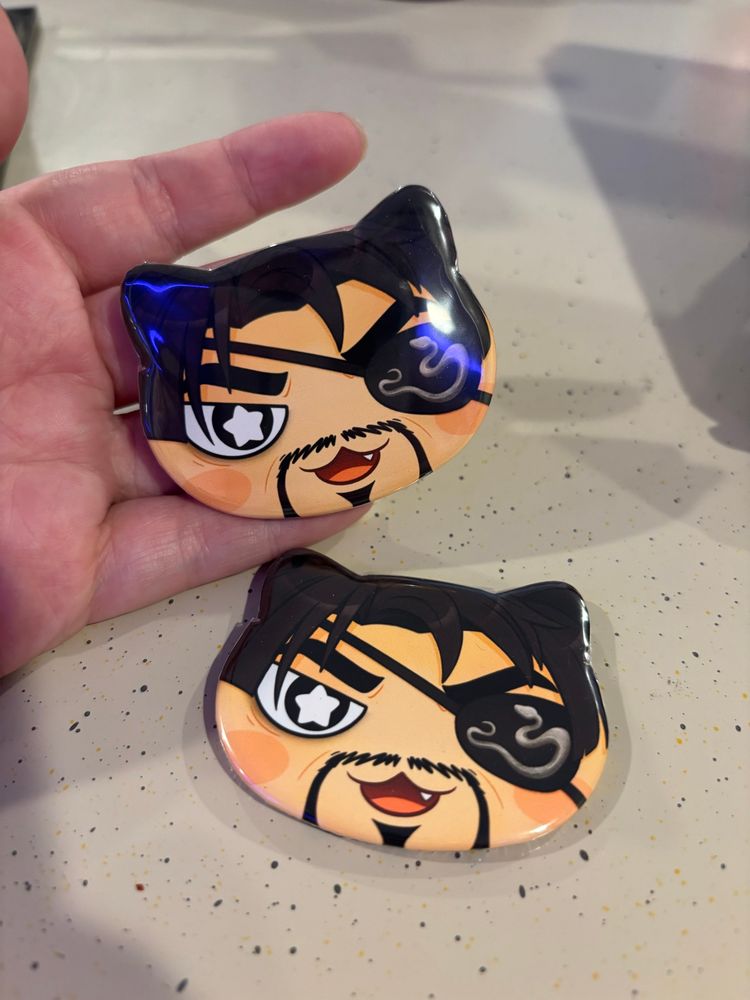 My hand holding two cat head-shaped buttons that are Majima’s face.