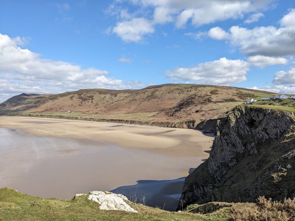 A photo of a wide sandy beach from the top of a cliff. There's hills behind the beach. The sky is blue with a few clouds