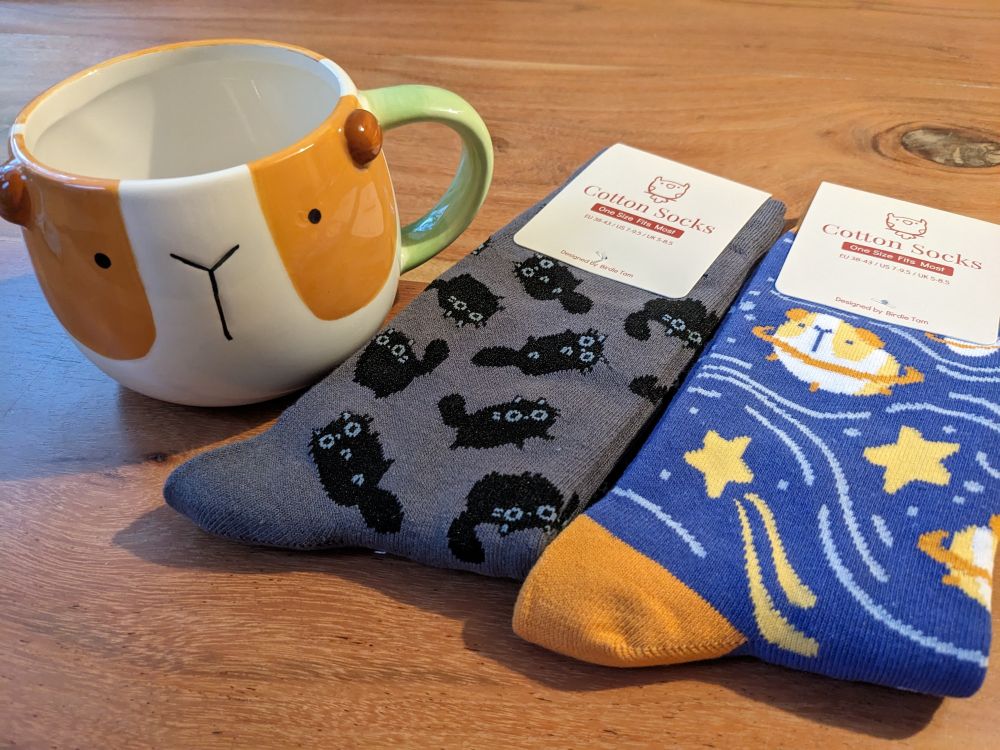 A mug painted like the head of a guinea pig with little knobs for ears, a pair of dark grey socks with black cats on them, and a pair of blue and orange socks with stars, planets and guinea pigs on them