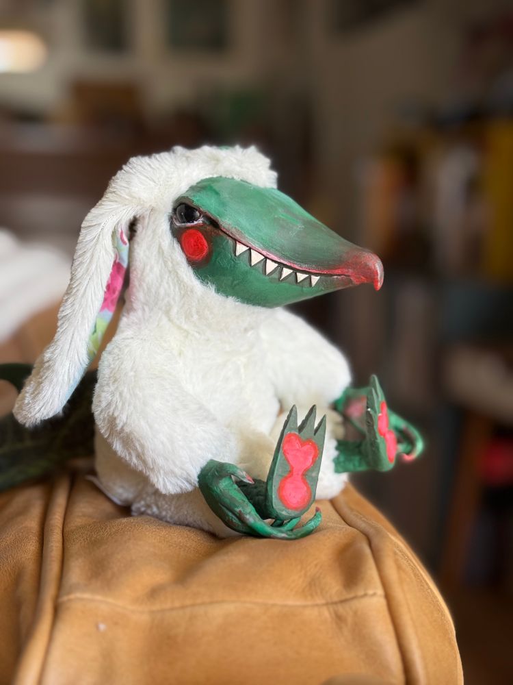 A custom doll with a lizard like face with a curved bird beak, sitting in a bunny suit