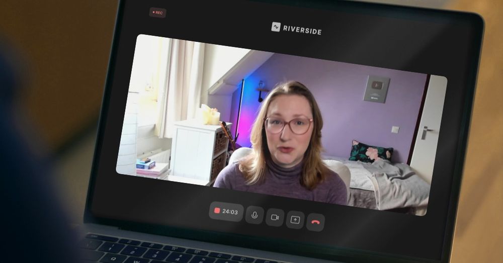 A screenshot of a podcast recording, a white woman making an awkward grimace, caught mid conversation, wearing a purple sweater and pink glasses.