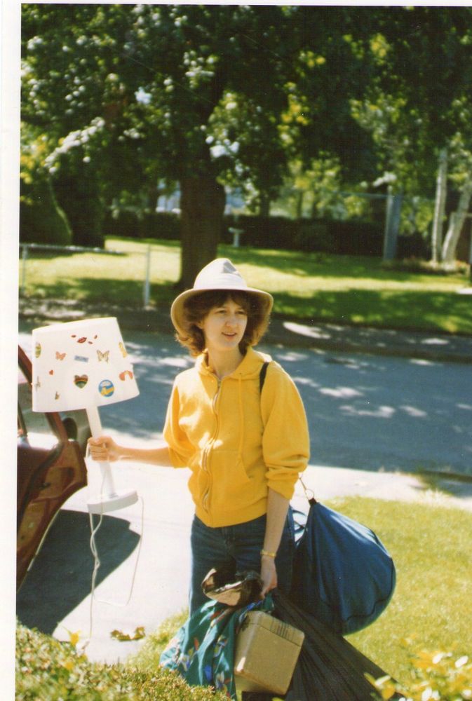 Me on a sunny day, wearing a tan fedora (?), yellow hoodie, and jeans, carrying several bags in one hand and a desk lamp in the other--the lampshade is festooned with many stickers, including several butterflies, a rainbow heart, and a cluster of red lips. My hair is feathered--it was the 80s. I'm not smiling, exactly, but there's a somewhat optimistic expression on my face.