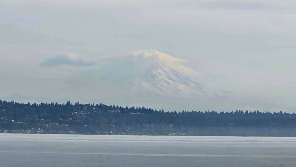 A distant view of Mount Tahoma (Mount Rainier) partly veiled by mist and clouds, with its snow-covered slopes faintly visible. In the foreground, Puget Sound stretches across the scene, bordered by a dark, tree-lined shoreline with scattered houses. The overall atmosphere is cool and hazy.

