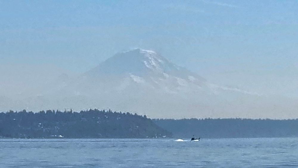 A hazy, distant view of a large snow-capped mountain rises behind a tree-covered shoreline. In the foreground, calm blue water stretches across the scene, with a small motorboat moving across it and leaving a white wake behind. The sky is clear and pale blue.