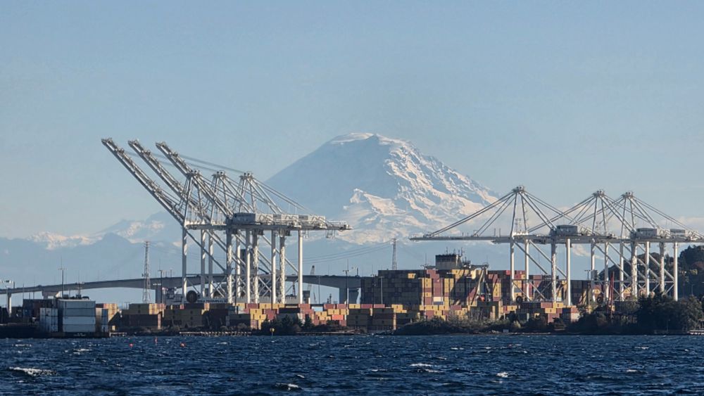 A view of Mount Tahoma (Mount Rainier) rising in the background behind the Port of Seattle. In the foreground, large white container cranes stand over stacks of colorful shipping containers along the waterfront, with the choppy blue waters of Elliott Bay in front. The snow-covered mountain dominates the skyline under a clear blue sky.
