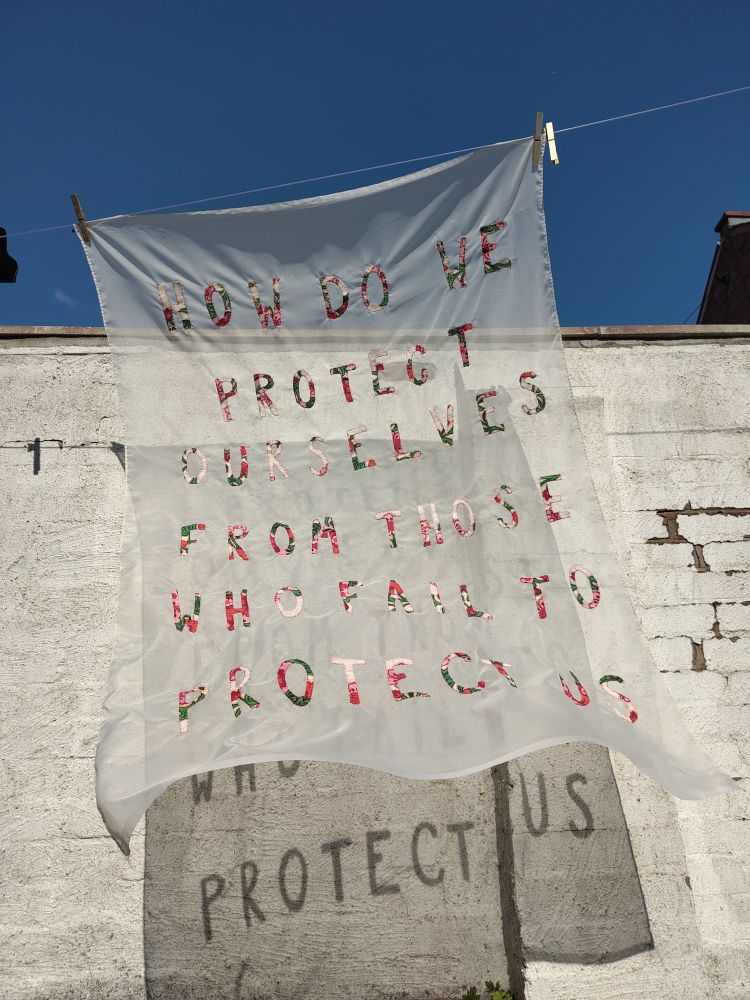 Photo of a sheer fabric artwork hanging from a washing line with a white wall and blue sky in the background.

The artwork is a sheer white fabric with text stitched in red and green fabric. The text reads: HOW DO WE PROTECT OURSELVES FROM THOSE WHO FAIL TO PROTECT US. As the fabric is sheer, the text can be read clearly in the shadow of the artwork. In this image the words PROTECT US are very clear in the shadow..